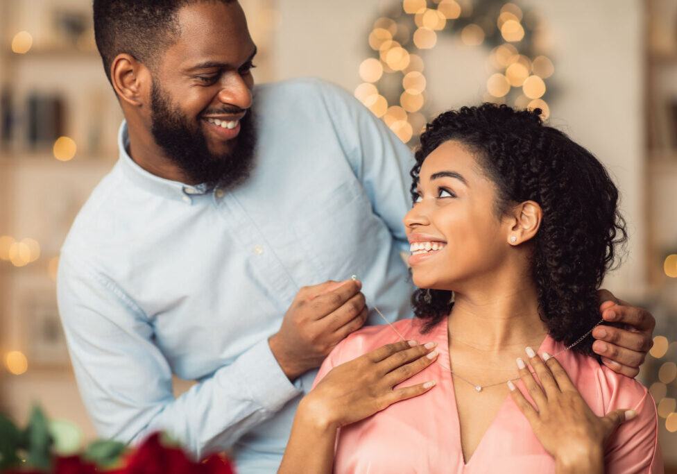 Couple smiling and exchanging jewelry, festive background.